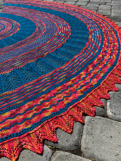 The All In Shawl by Destiny Itano, A picture of a half circle two-color shawl with multiple stitch patterns and a zig-zag edge. Colors are blue and a variegated reds/oranges/pinks.
