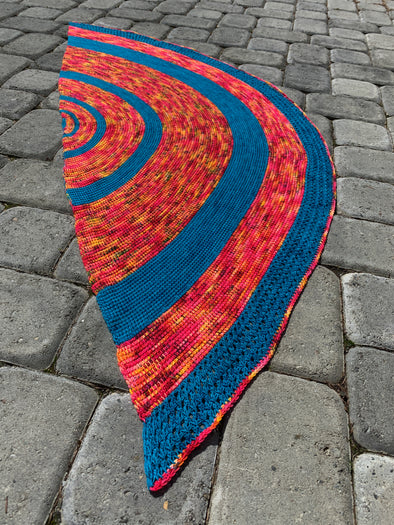 A picture of a half circle Tunisian crochet shawl, with large, alternating bands of two colors, laying on grey stones. Colors are blue and a variegated reds/oranges/pinks.