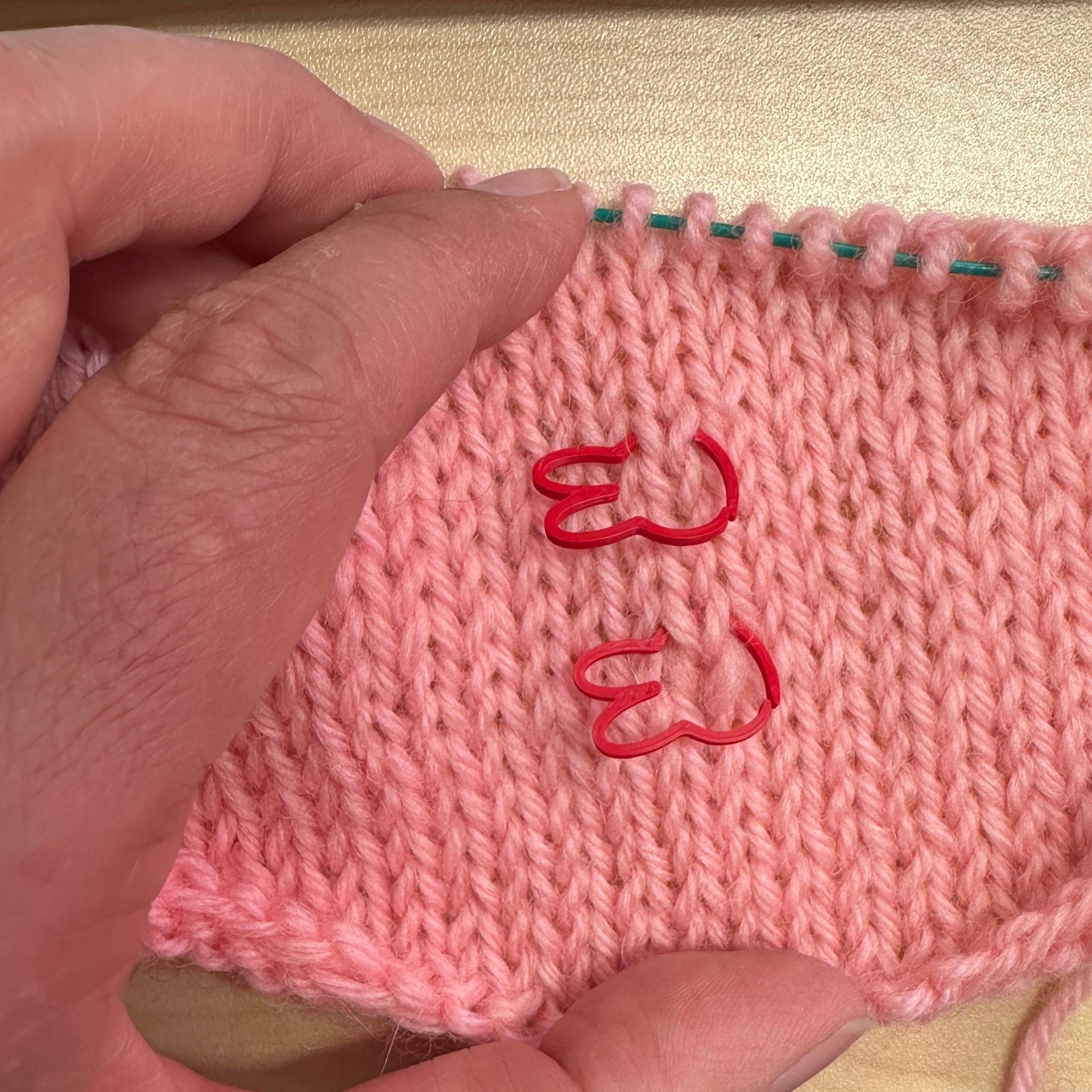 A close-up of a hand holding a knitted pink fabric swatch with red rabbit-shaped stitch markers on the knitting. 
